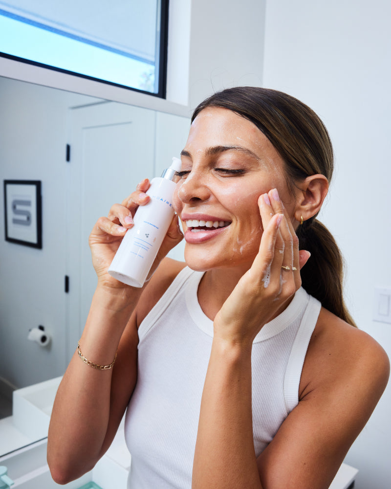 Smiling woman applying Bio-Cleanse 5.5  Face wash lather to face in bright bathroom gallery position 1
