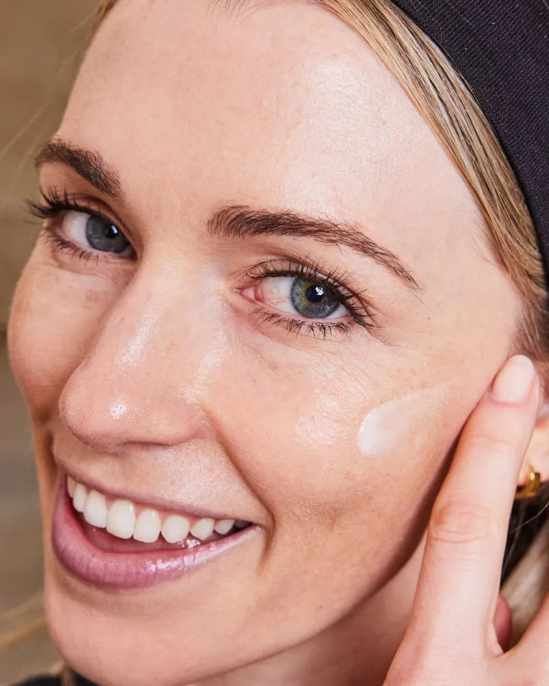 Woman smiling while applying Daily Skincare Bundle cream to cheek in g