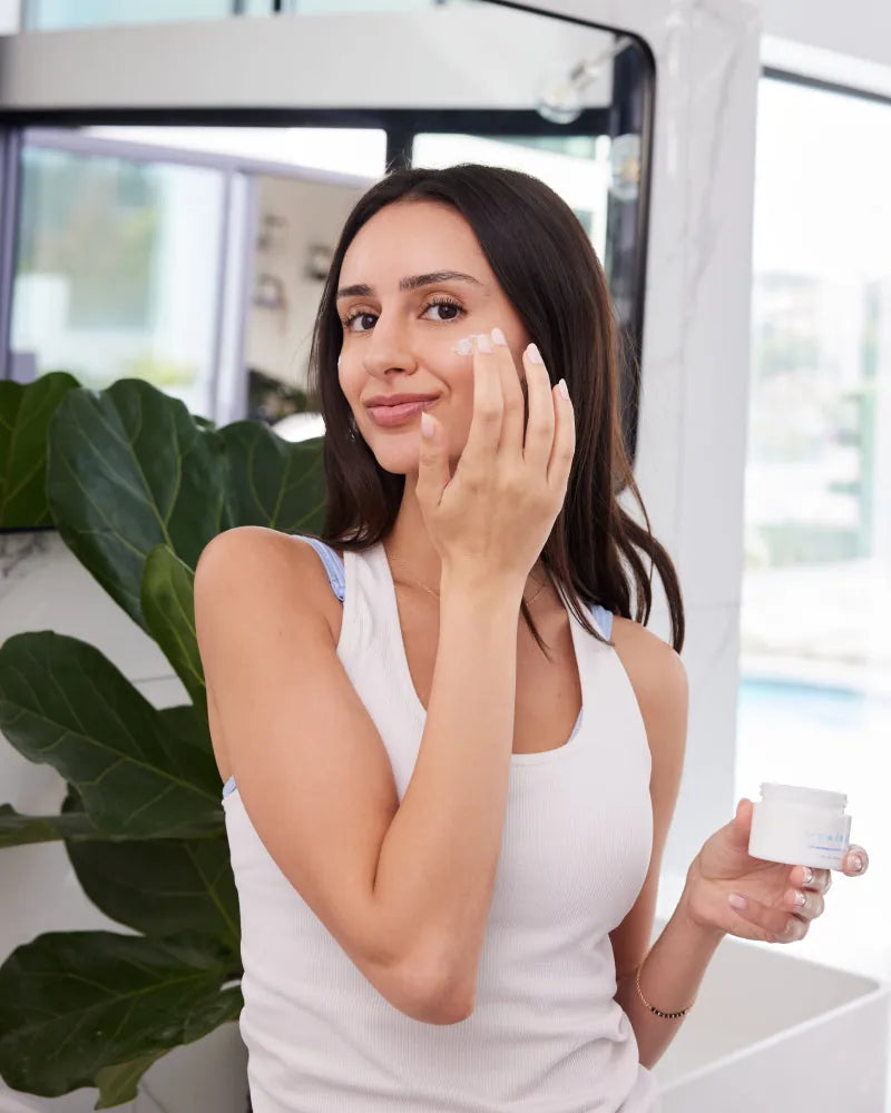 Woman applying NMF Intense Barrier Cream on cheek holding jar indoors 